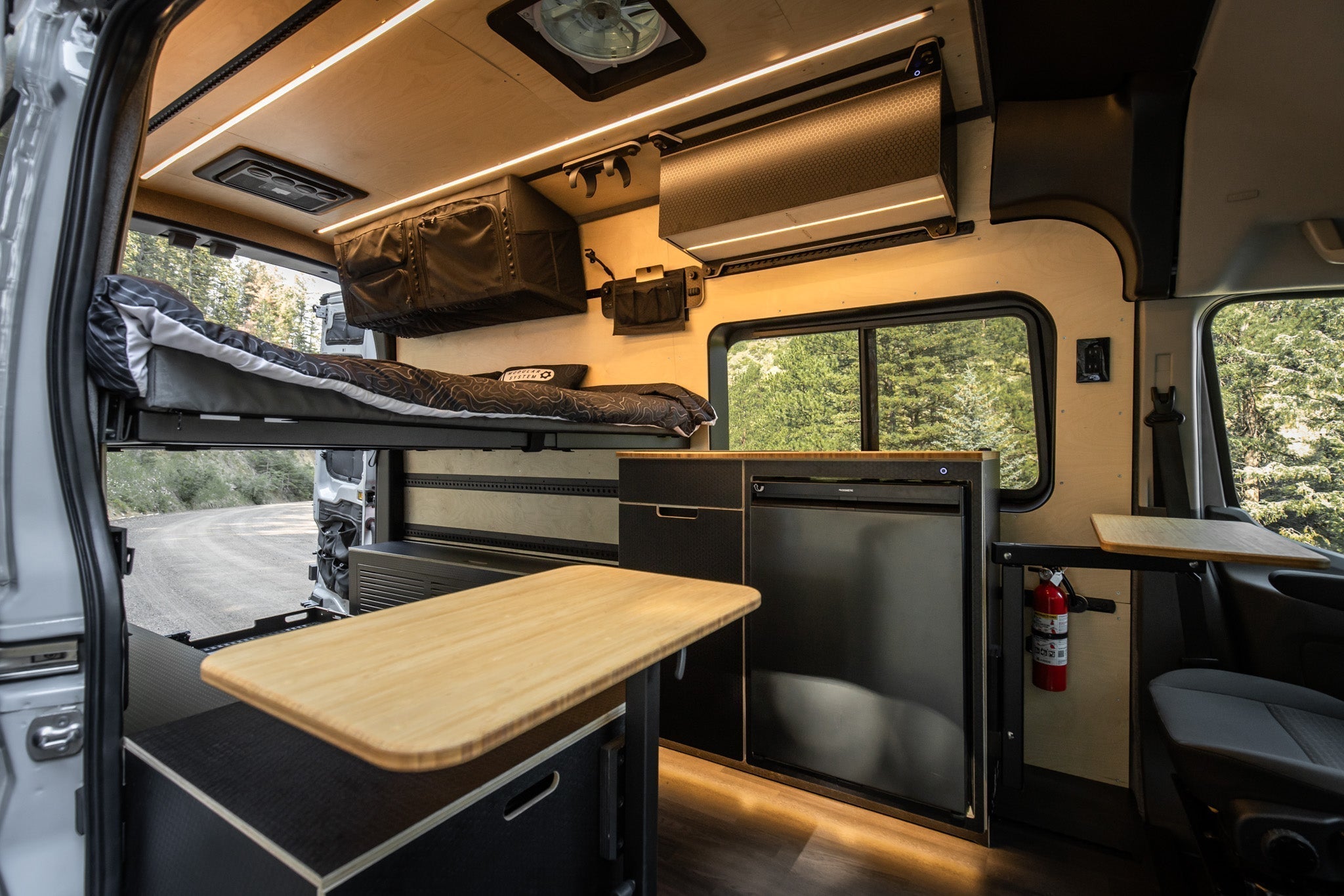 Interior of a modern camper van with a neatly made bed above a wooden table. Kitchenette with fridge and storage, alongside large window and forest view. Cozy and efficient.