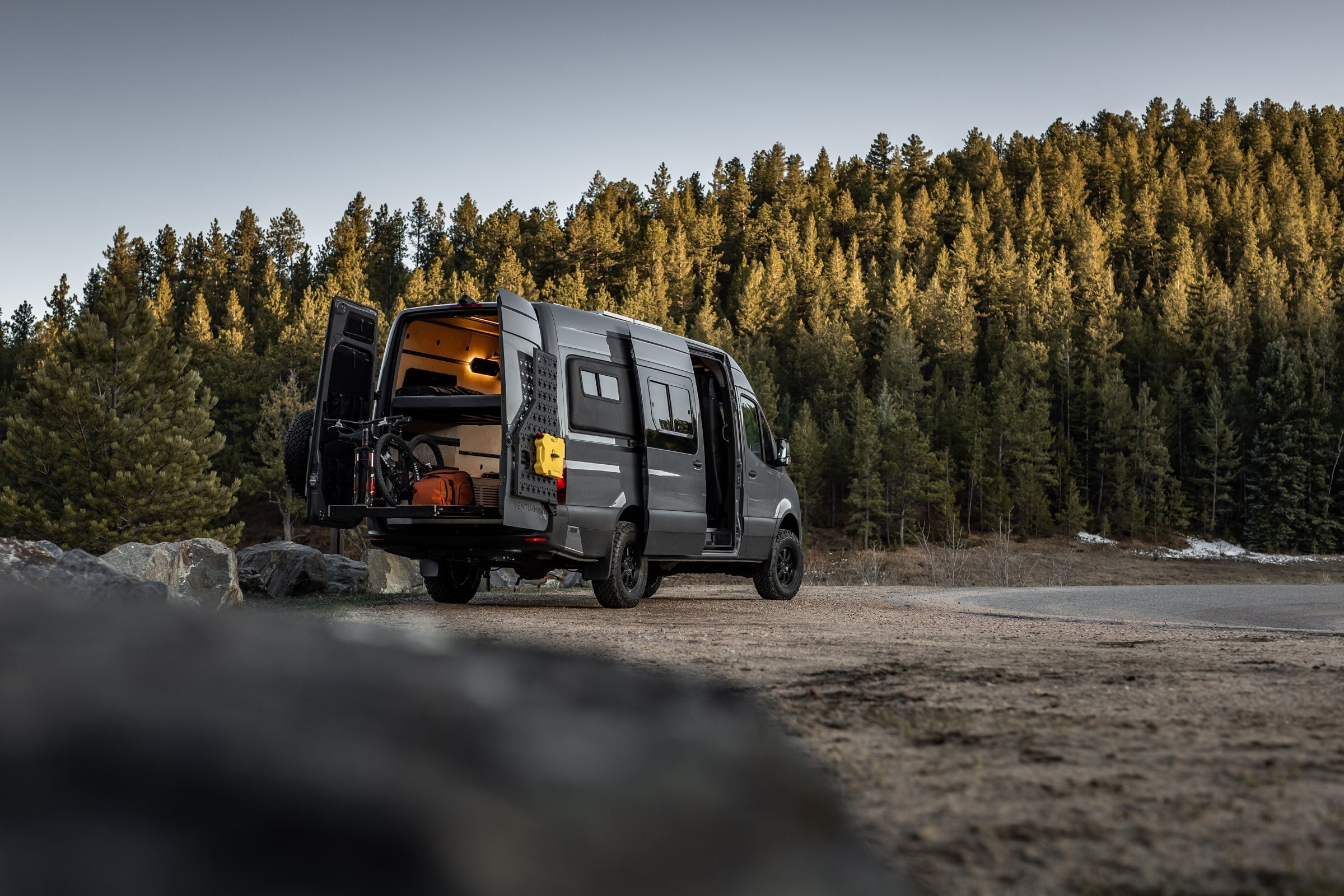 A black camper van with its rear doors open is parked on a dirt area near a forest of tall pine trees in daylight.