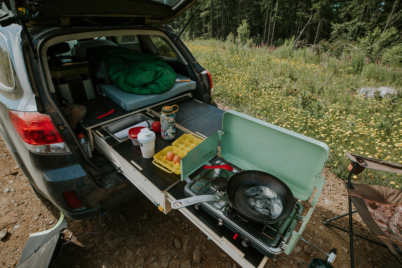 Open car trunk with the Trekhaus SUV Camper kit drawers open with camping gear and a portable stove outdoors